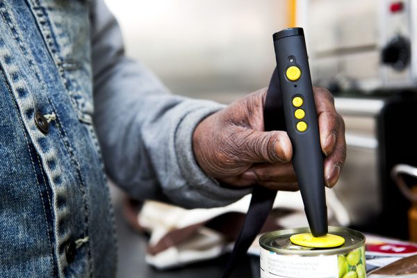 un lecteur vocal d'étiquettes tenu en main par une personne et l'embout posé sur une étiquette magnétique placée sur une boite de conserve. Sur la face devant du lecteur on voit trois petits boutons et une grosse